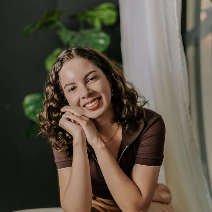 A psicóloga Barbara Domingos, de cabelo cacheado, sorri para a câmera, apoiando o rosto nas mãos entrelaçadas. Ela está vestindo uma blusa polo marrom e calças de cor bege. Atrás dela, uma parede escura e uma planta grande de folhas verdes.